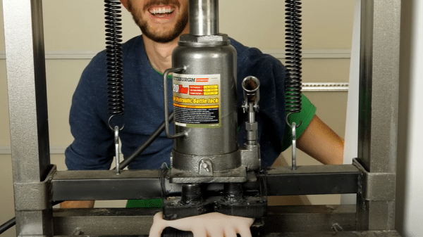 Featured image. Man grins as a rubber hand he believes to be his own is crushed in a hydraulic press. Credit to The Action Lab on YouTube.