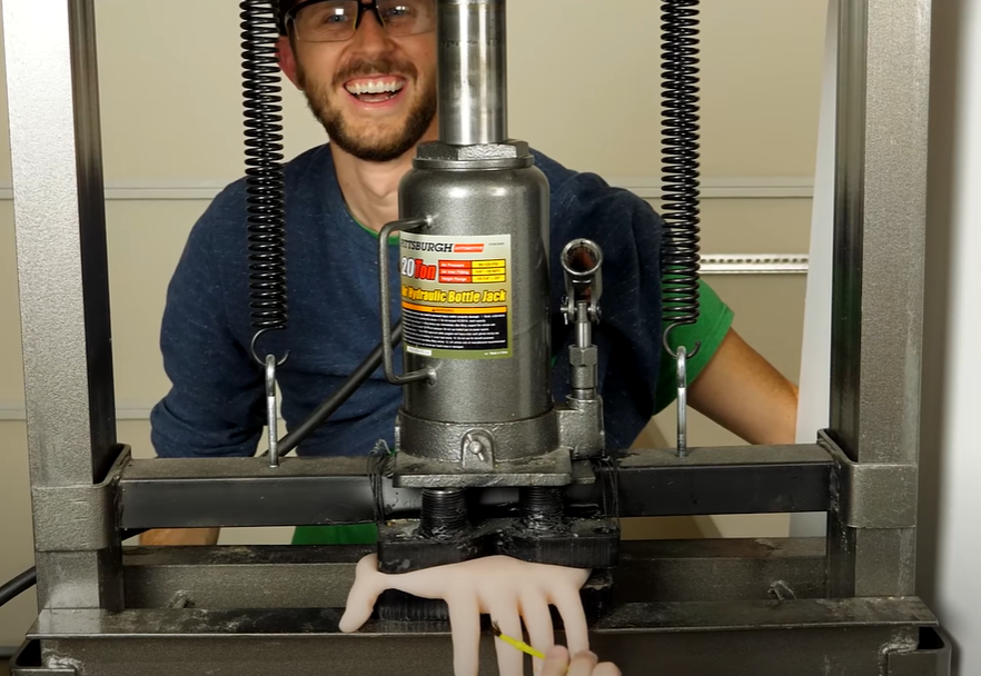 Featured image. Man grins as a rubber hand he believes to be his own is crushed in a hydraulic press. Credit to The Action Lab on YouTube.