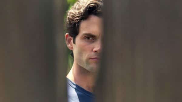 Credit to Netflix. Joe Goldberg looks through the hole of a fence.