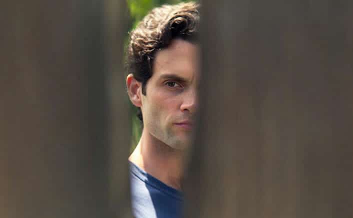 Credit to Netflix. Joe Goldberg looks through the hole of a fence.