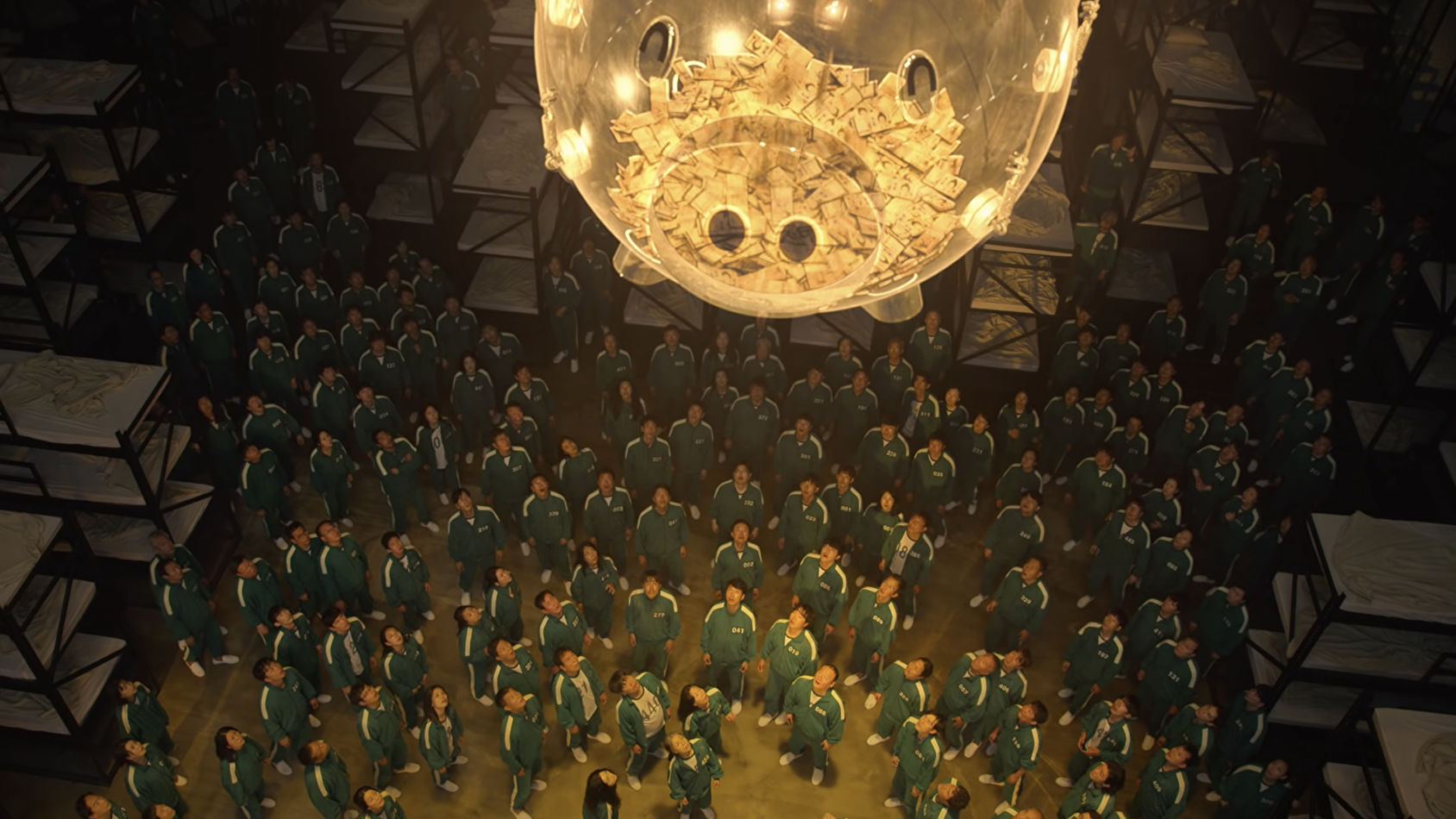 Credit to Netflix. Squid Game players look up at the big golden piggy bank full of money.