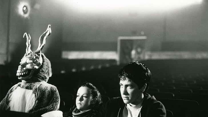 A bunny-suited man, a young woman, and young man sit side-by-side in an empty theater.
