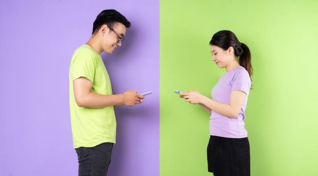 Asian couple texting each other and smiling against bright green and purple backgrounds