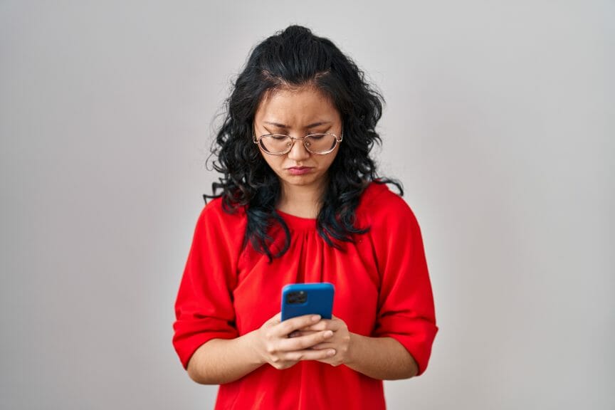 Asian woman with glasses looking at her phone with an upset expression