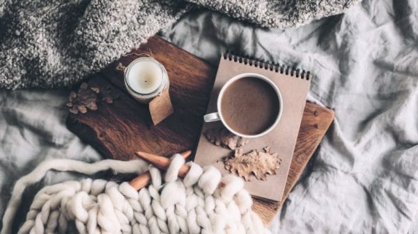 Candle, hot drink, notebook, and knitting on a cozy bedspread