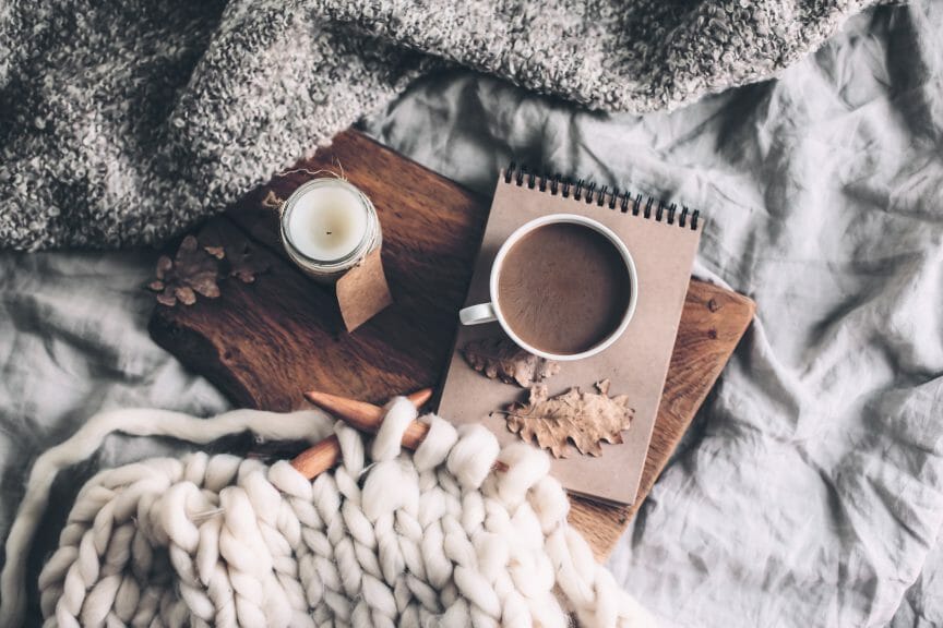 Candle, hot drink, notebook, and knitting on a cozy bedspread