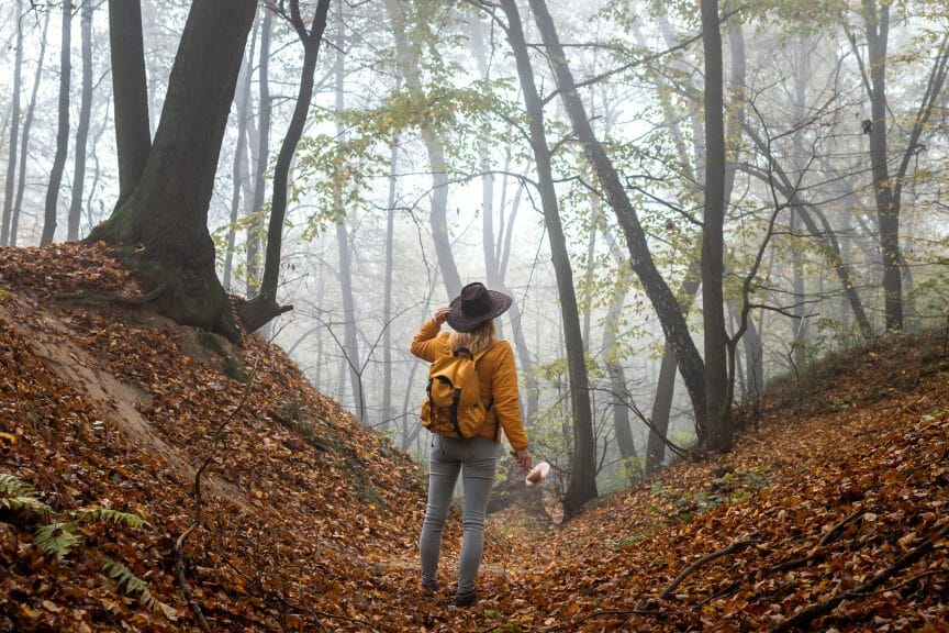Woman wearing a wide-brimmed hat a mustard yellow jacket and backpack standing in the woods with fallen leaves