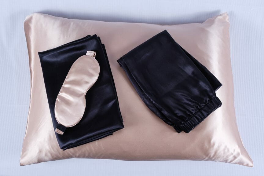 Soft pink silk pillow with black sleep set sitting on top and pink silk sleep mask.