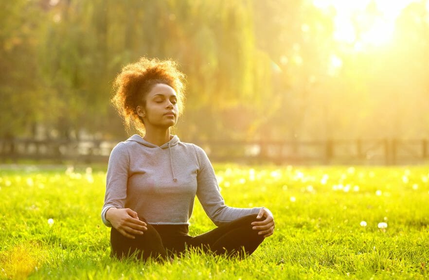Woman sitting in the grass with her eyes closed for mind-body connection.