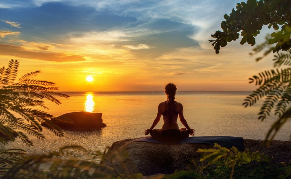 Woman meditating on the shoreline sitting on a rock for mind-body connection.