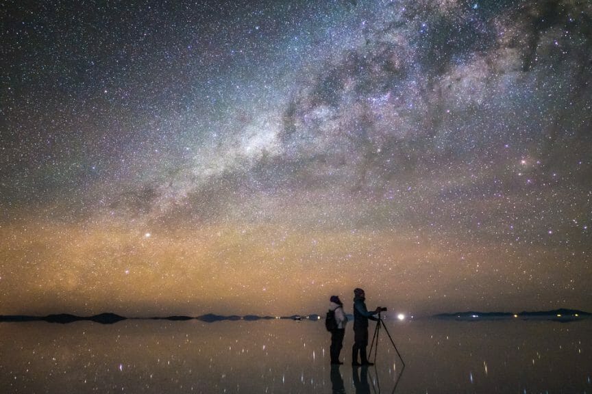 Two people standing on a salt flat looking up at all of the stars.