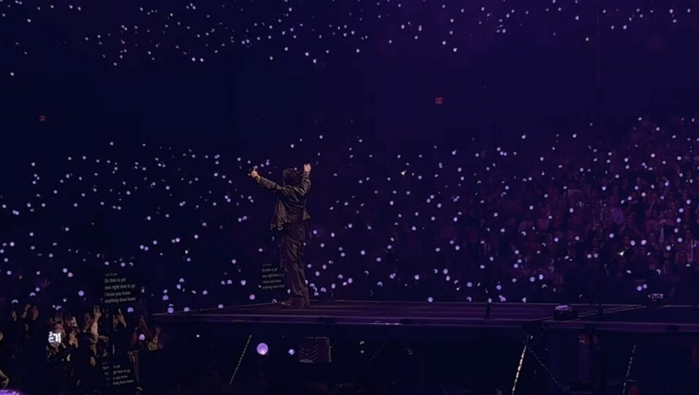 Agust D throwing his hands in the air in front of a sea of purple light sticks. 