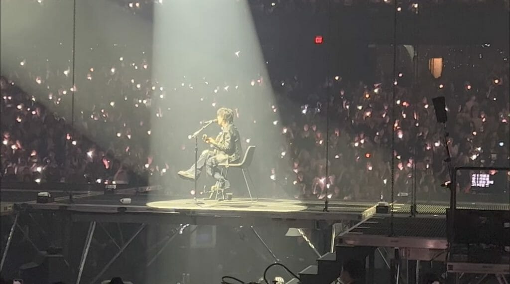 Agust D sitting on a chair playing the guitar.