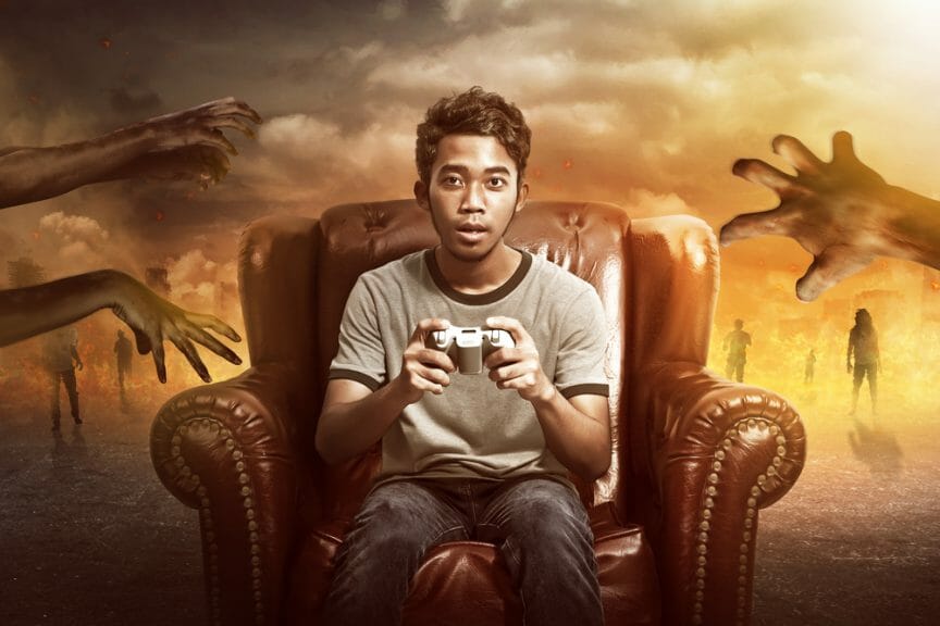 Man sitting on sofa with gaming remote in hand with zombie hands reaching towards him in the background.