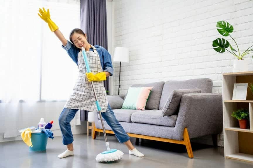 A woman cleaning her house, dancing at the same time. She is holding a mop and has one hand in the air.