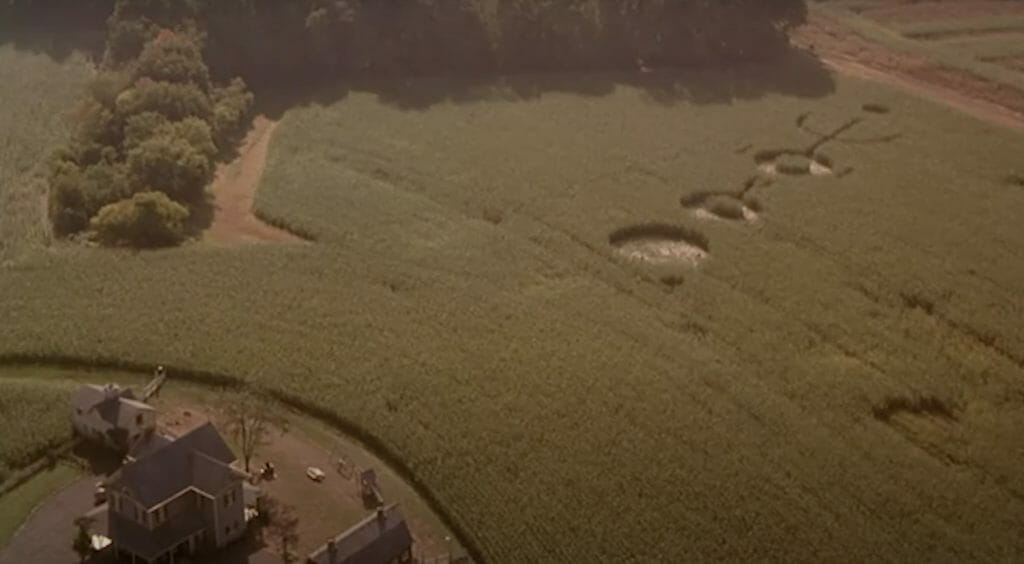 An aerial view of green crops with a farmhouse at the bottom left, and three large crop circles done by aliens.