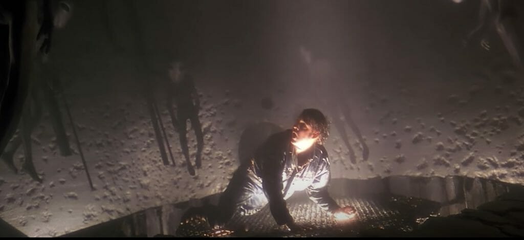 A man, Travis Walton, is on all fours looking up with a horrified expression at alien suits that seem to be staring down at him.