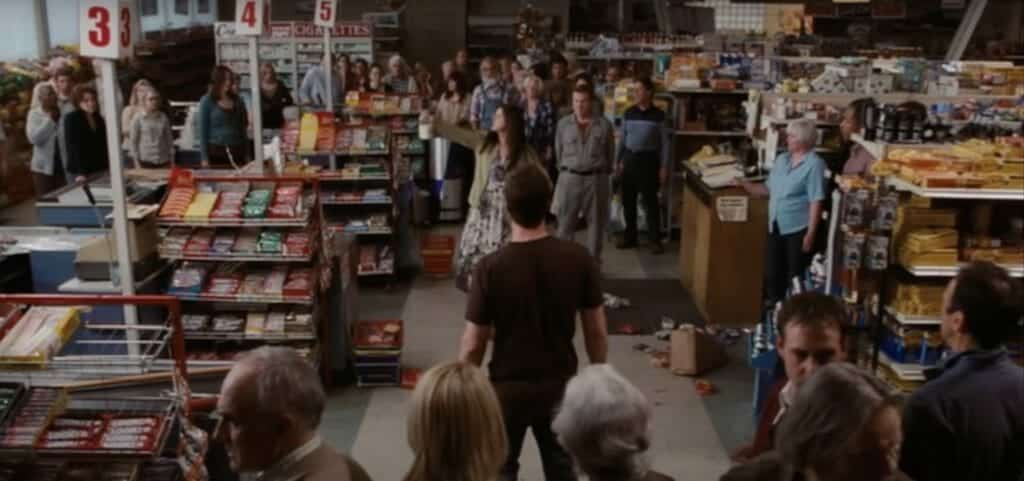 Several people stand inside a market, with one group of people being cornered by another larger one. A Woman points outside the market windows to the mist and its alien creatures.
