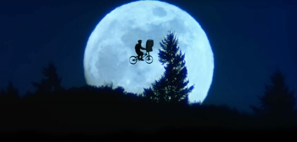 The silhouette of a boy and an alien on a bike flying in front of a big round moon and over a dark forest. 