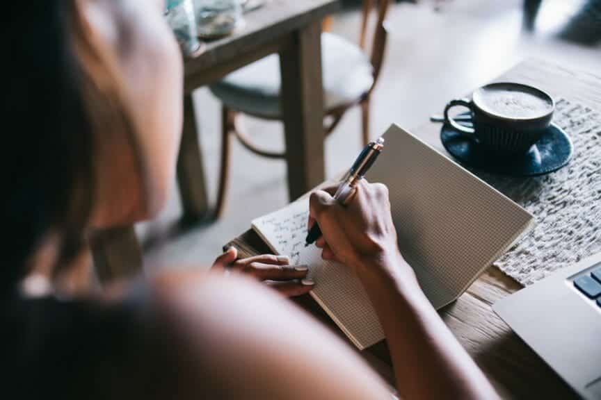 Back view of crop anonymous woman writing notes in notebook with pen while sitting in room with cup of coffee.