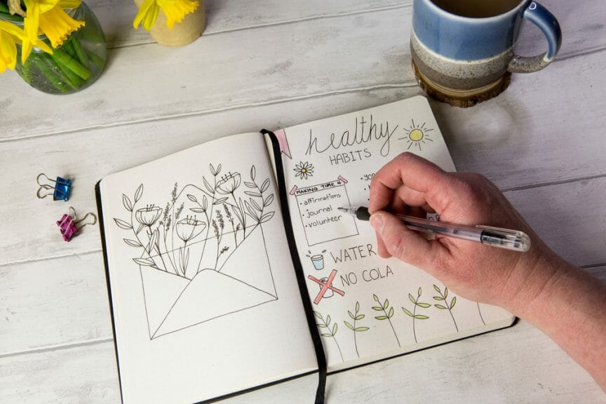 Bullet journal open on self care healthy habits layout pages with hand holding pen. Over shoulder view, fresh white table background, flowers, positive mental health message.