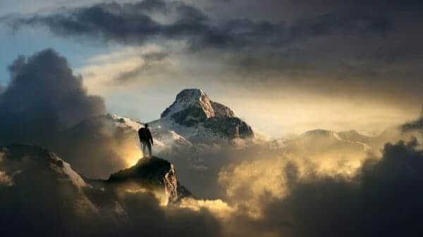 Image of a climber standing atop a mountain, looking into the distance where another mountain stands.