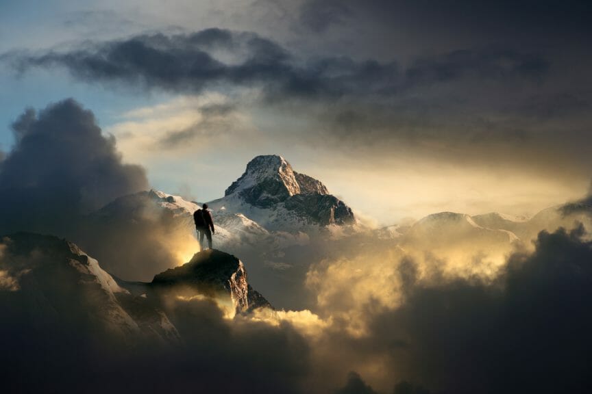 Image of a climber standing atop a mountain, looking into the distance where another mountain stands.