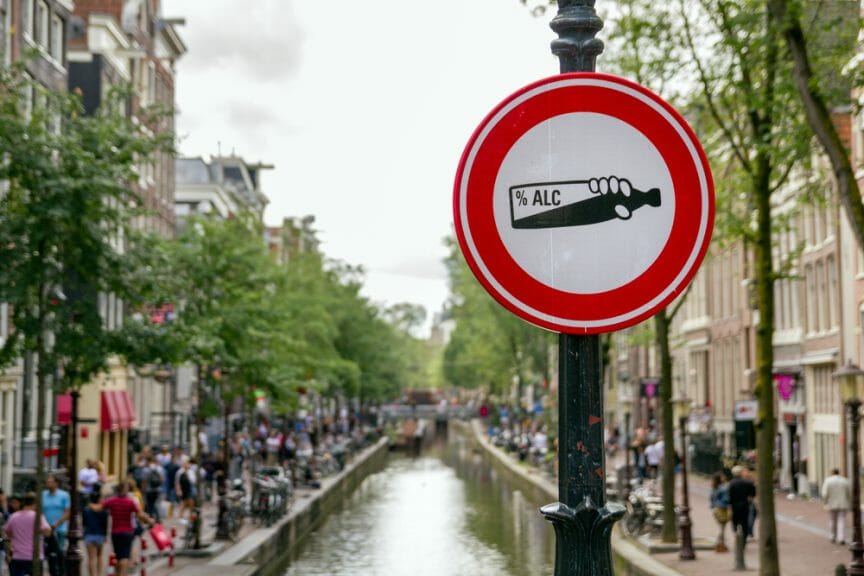 A red street sign by the Amsterdam canal. It displays an empty alcohol bottle to signal the 'no-alcohol zone'.