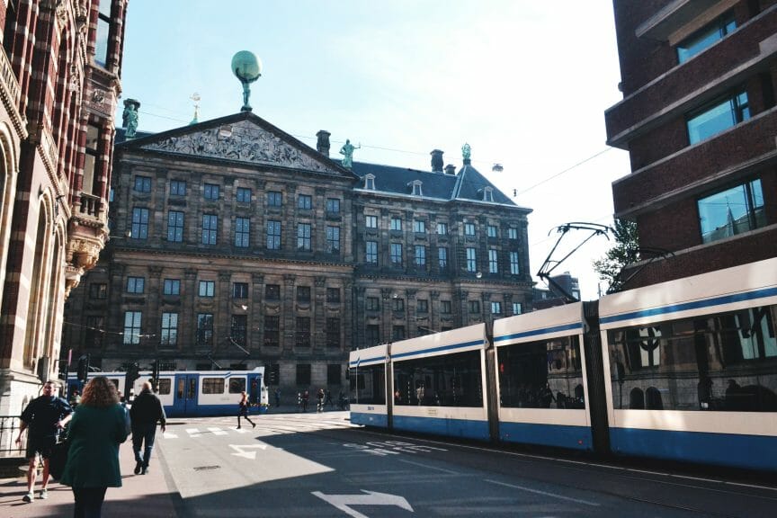 A busy Amsterdam street. People walking and a tram can be seen in front of a large building. 