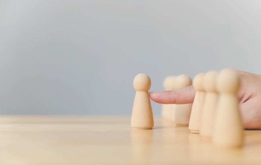 Plain, small figurines placed in a horizontal row with a hand pushing one figurine outside of the row.