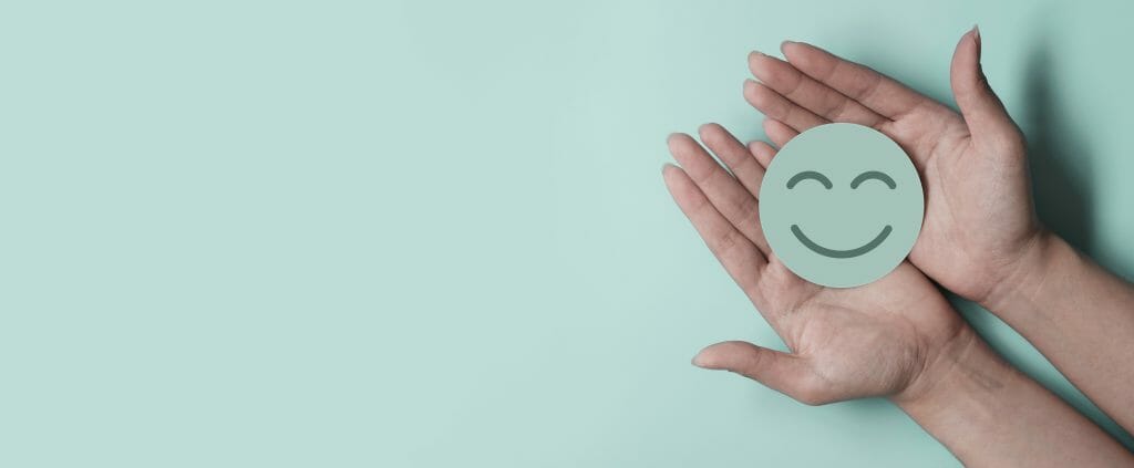 This image has a turquoise background with two cupped hands on the far-right side of the picture holding a turquoise smiley face. 