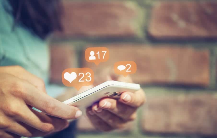 An image of someone using social media on their phone with 'comments,' 'likes,' and 'friends' in three separate small dialogue bubbles.