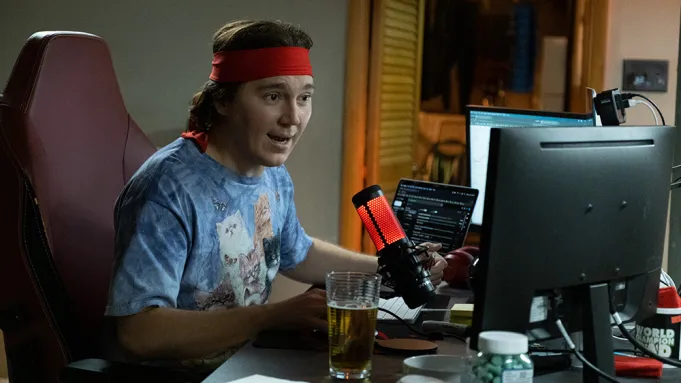 Paul Dano in 'Dumb Money' speaking into a microphone and facing a set of computers. 