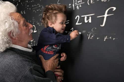 Image of a man resembling Einstein holding up a child to complete an equation on a blackboard.