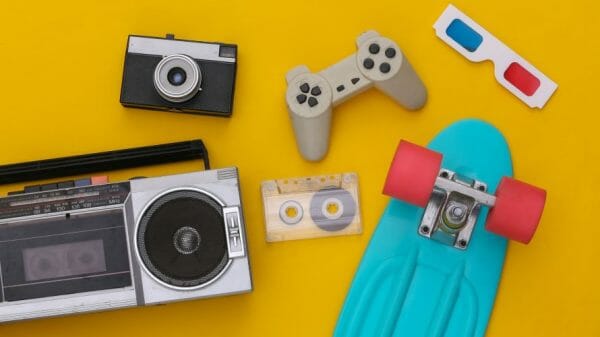 1980's items lay on a yellow background. They include a camera, game console, 3D glasses, a skateboard, a stereo, and a cassette tape.