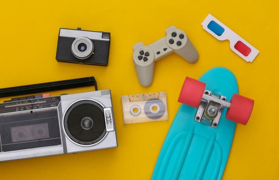 1980's items lay on a yellow background. They include a camera, game console, 3D glasses, a skateboard, a stereo, and a cassette tape.