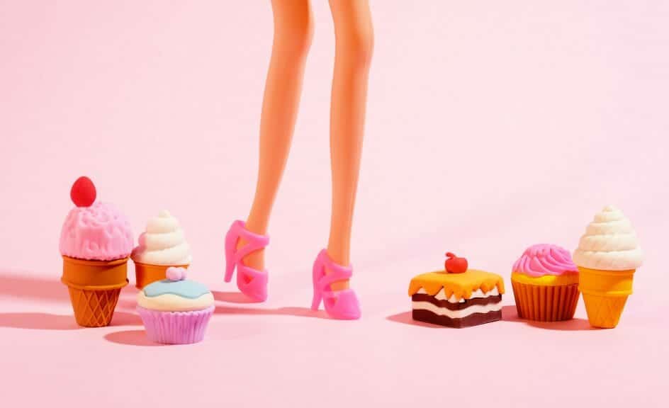 A pink background with only the Barbie's legs in the frame. She's wearing pink heels and has treats next to her feet.