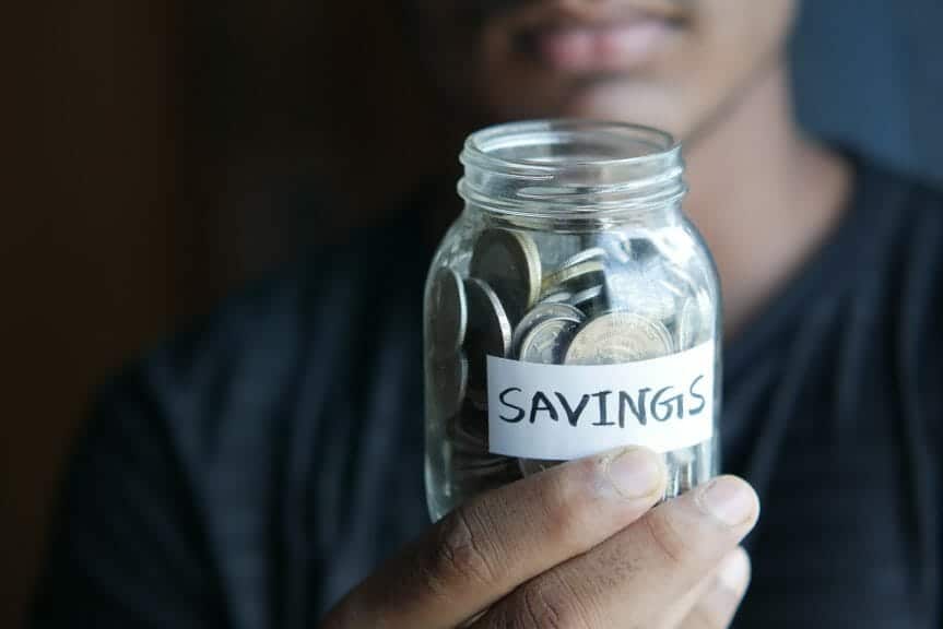 A man is holding a jar labelled 'Savings'. The jar is filled with coins.