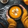 Female hands in yellow knitted sweater holding a bowl with pumpkin cream soup on dark stone background with spoon decorated with cut fresh pumpkin, top view.