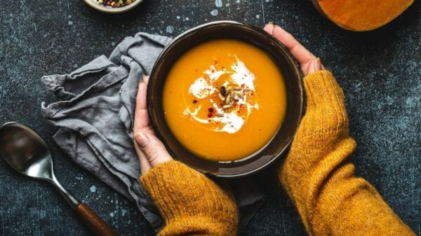 Female hands in yellow knitted sweater holding a bowl with pumpkin cream soup on dark stone background with spoon decorated with cut fresh pumpkin, top view.