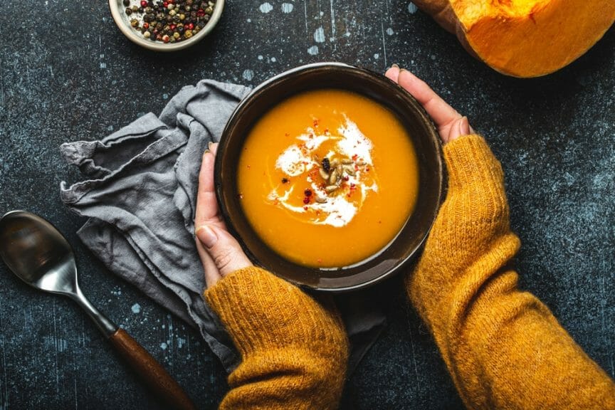 Female hands in yellow knitted sweater holding a bowl with pumpkin cream soup on dark stone background with spoon decorated with cut fresh pumpkin, top view.