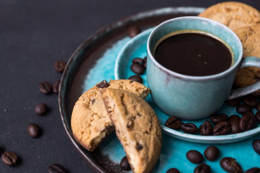 A cup of coffee won a blue plate with pieces of chocolate-chip cookies.