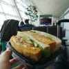 someone is holding a sandwich in the aiport, the airport window can be seen on the lefthand side as well as a suitcase handle in the right corner of the photo