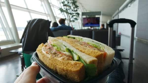 someone is holding a sandwich in the aiport, the airport window can be seen on the lefthand side as well as a suitcase handle in the right corner of the photo