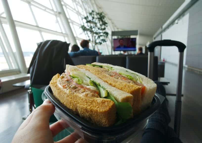 someone is holding a sandwich in the aiport, the airport window can be seen on the lefthand side as well as a suitcase handle in the right corner of the photo