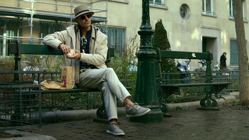 Michael Fassbender sitting on a park bench and eating McDonald's in a scene from 'The Killer.'