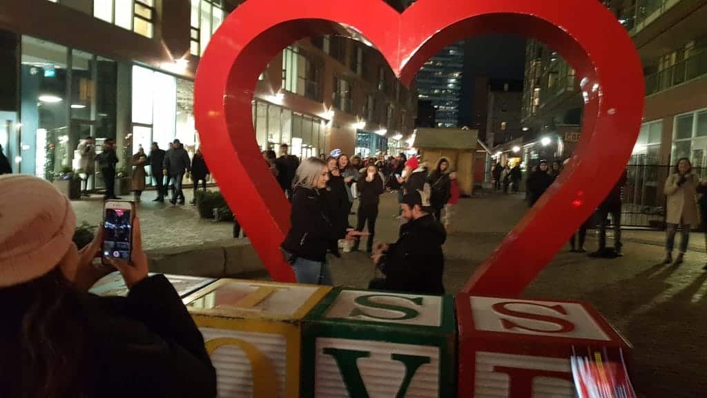 A man proposing to a woman under a heart-shaped structure at night with the word "love" in block letters under them and people in the background.