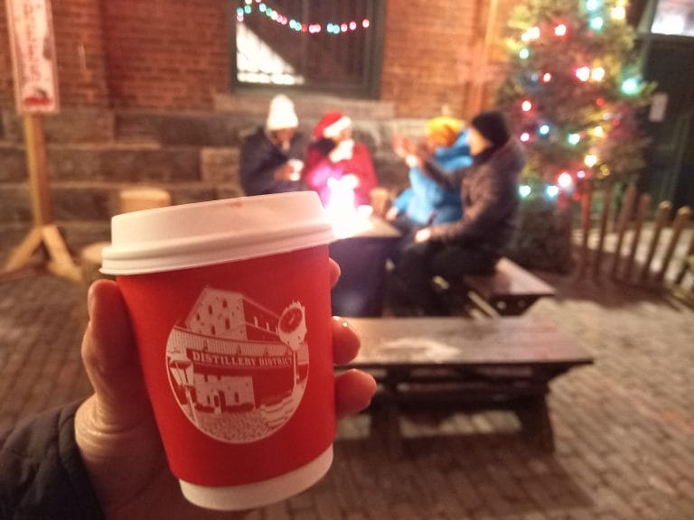 A red and white up of hot chocolate with people sitting and a Christmas tree in the background.