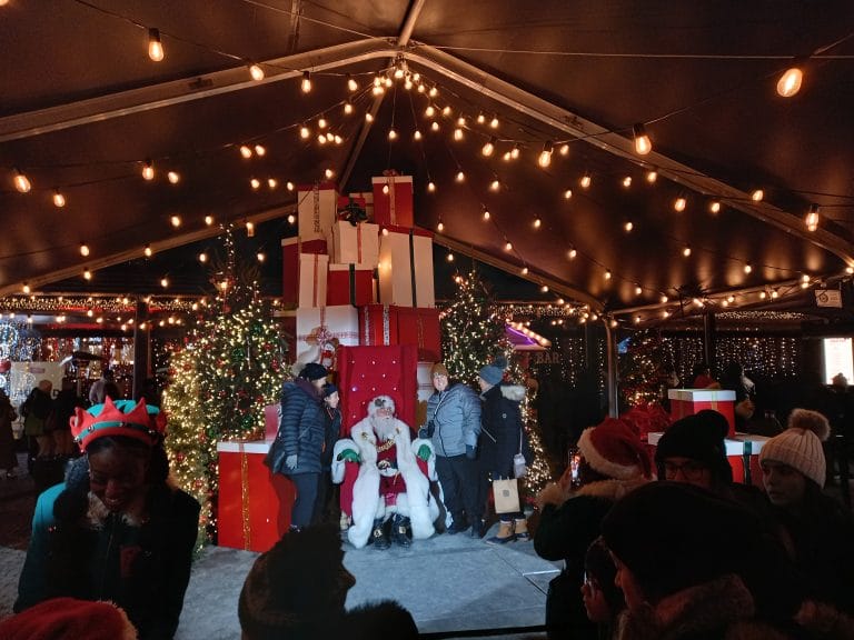 Under a canopy with string lights, a thrown with presents behind it and Christmas trees beside it hold Santa, taking photos with people.