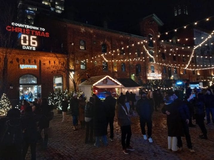 A crowd of people underneath lights strung between buildings. A countdown to Christmas is above one of the buildings, displaying 6 days left.